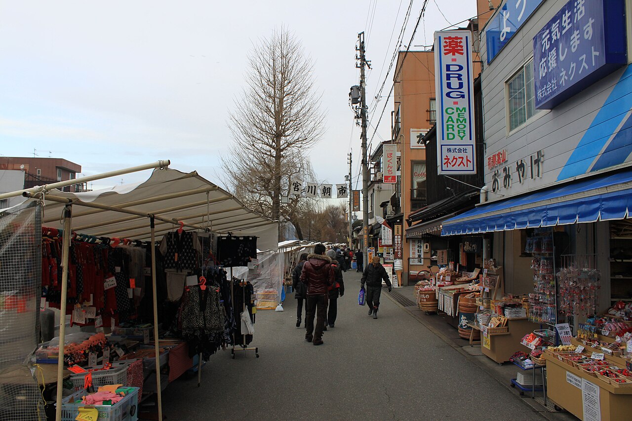 Market stalls with canvas awnings lining a street at the Miyagawa Morning Market in Takayama, shoppers walking past small shops under a bare ginkgo tree.
