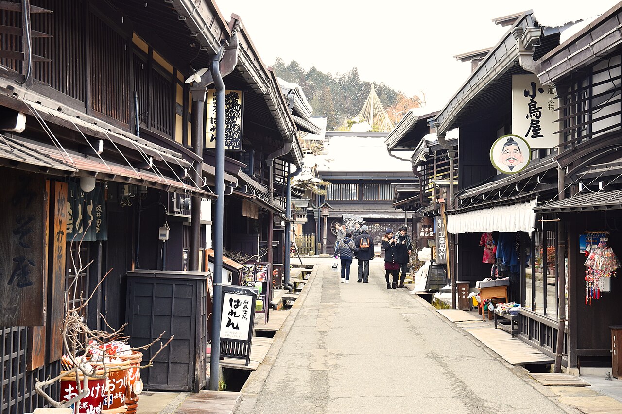 A narrow street of dark wooden merchant houses with latticed fronts and shop signs in the Sanmachi Suji old town of Takayama, a few visitors walking ahead.
