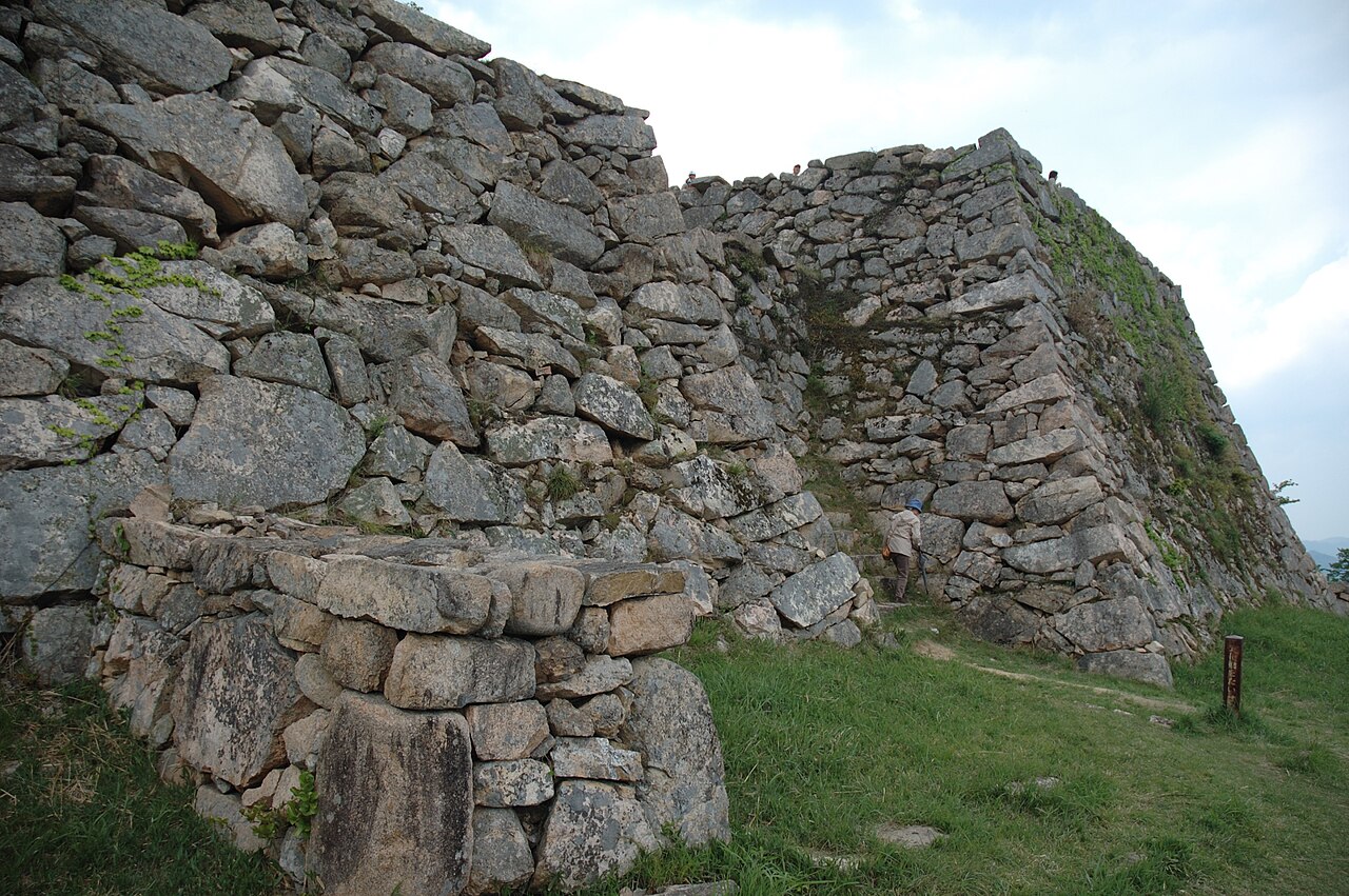 Close view of rough-hewn stone walls at the Takeda Castle ruins, with grass in the foreground and a visitor near the base of the wall.