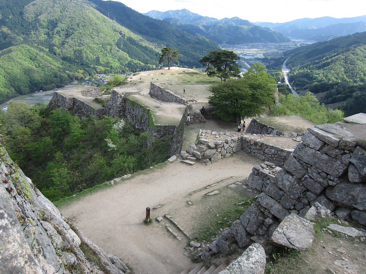 Stone walls and flat terraces of the Takeda Castle ruins on a mountaintop, with green forested ridges and a valley below.