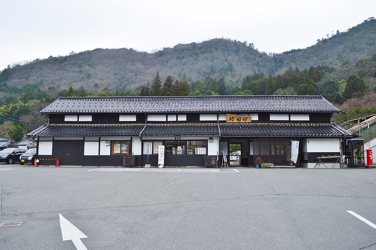 Front of Takeda Station, a long low building with a dark tiled roof and white walls, across an empty parking area with a wooded hill behind.