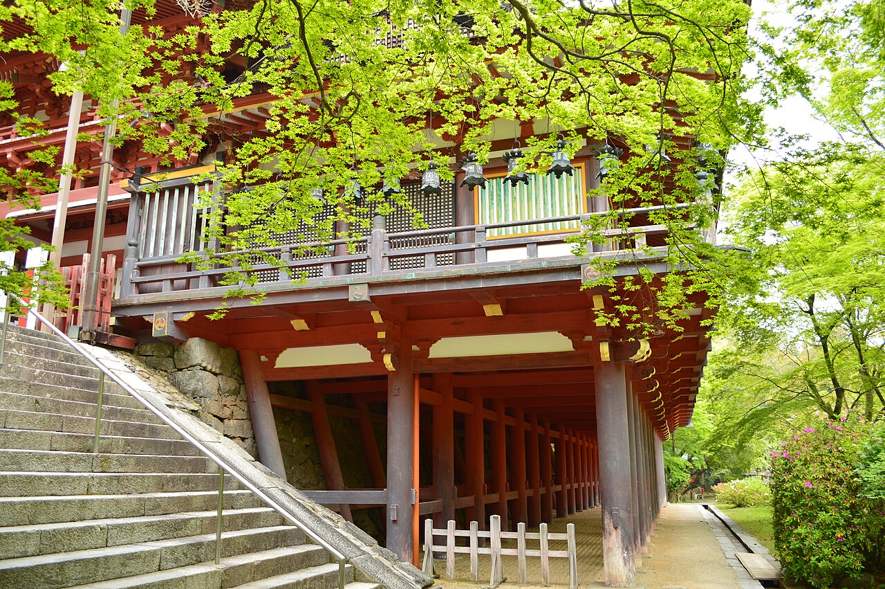 A red wooden hall stands on tall pillars above a broad stone staircase, with lanterns hanging along its balcony under fresh green maple leaves