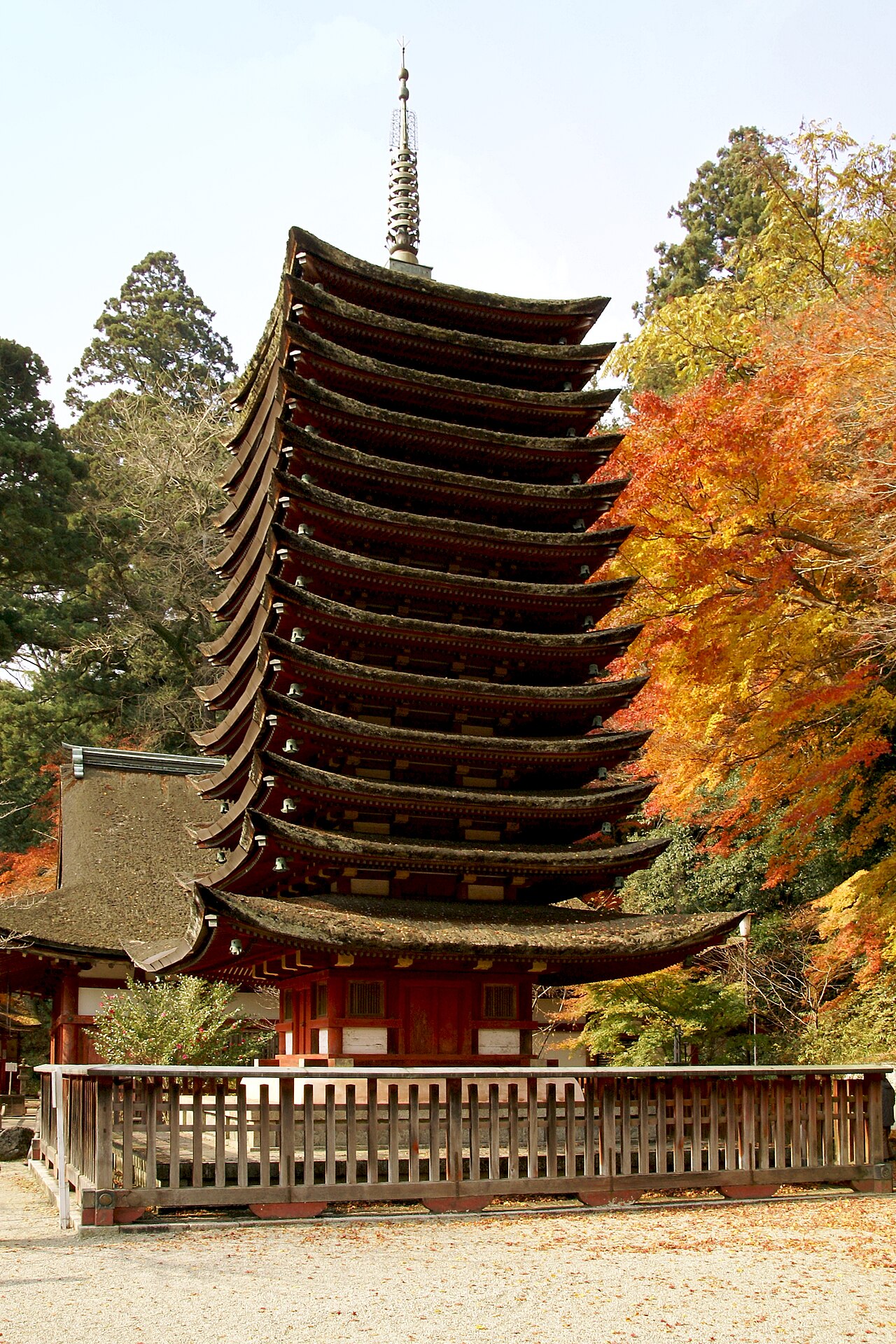 A tall thirteen-tier wooden pagoda with moss-dark roofs stands behind a low wooden fence, with orange and yellow maple leaves on the right