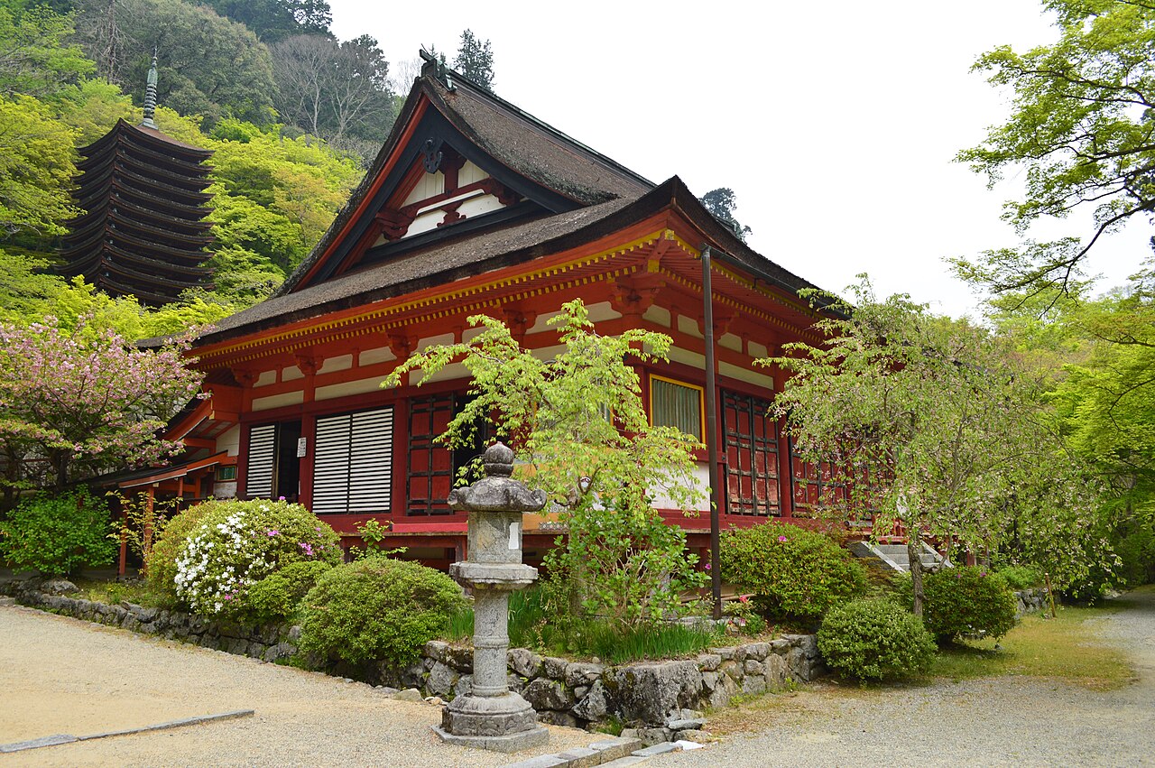A red and white wooden worship hall with a gabled roof stands beside a stone lantern, with the pagoda showing above fresh green trees and azalea bushes