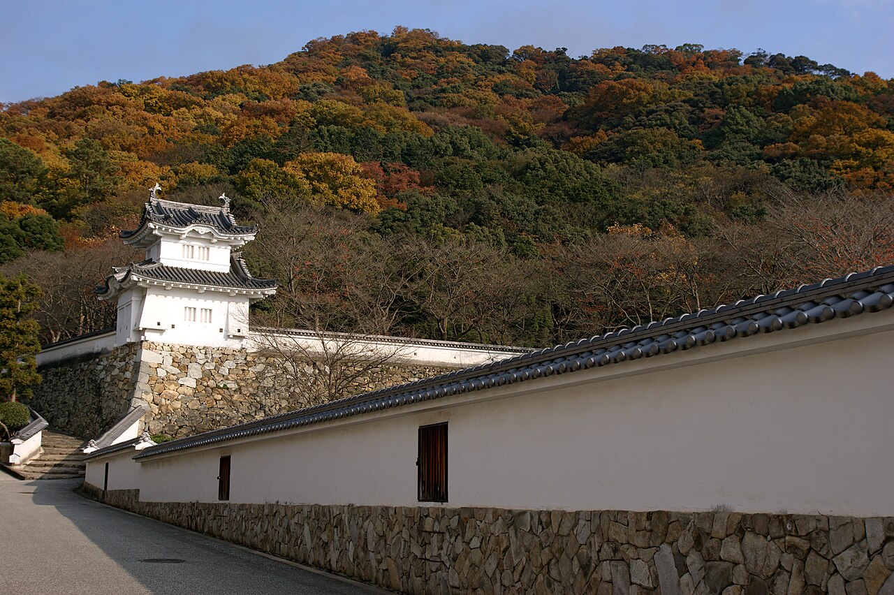 The corner turret of Tatsuno Castle above white walls and a stone foundation, with a wooded hillside of autumn colour behind.