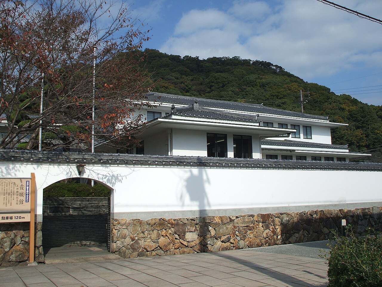 The white-walled, tile-roofed Kajokan in Tatsuno behind a long white wall with a stone base, with a wooded hill rising behind.