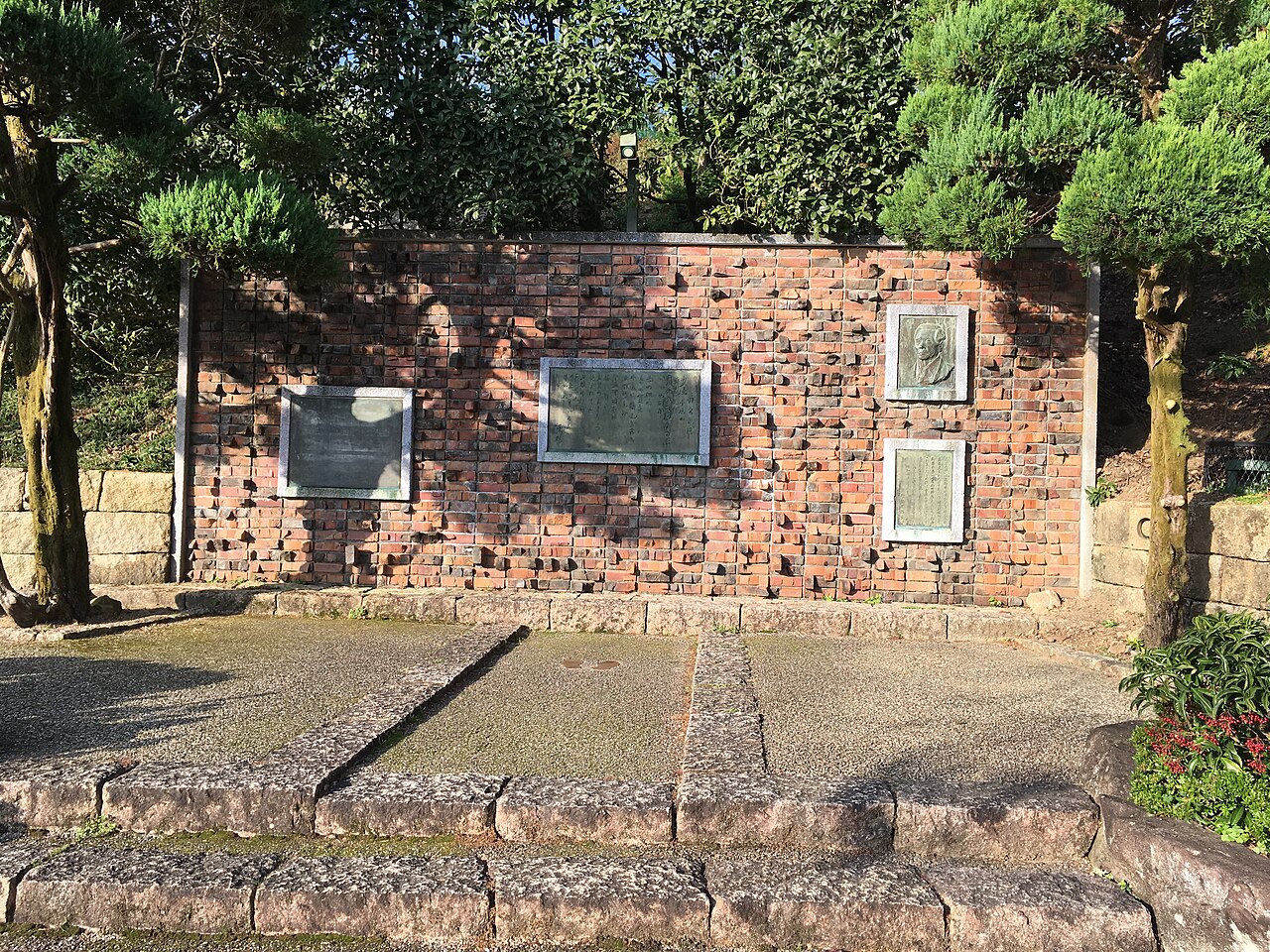Brick wall with bronze plaques and a portrait relief of the Akatombo (Red Dragonfly) song monument to poet Miki Rofu in Tatsuno, framed by trimmed pines and a stone step.