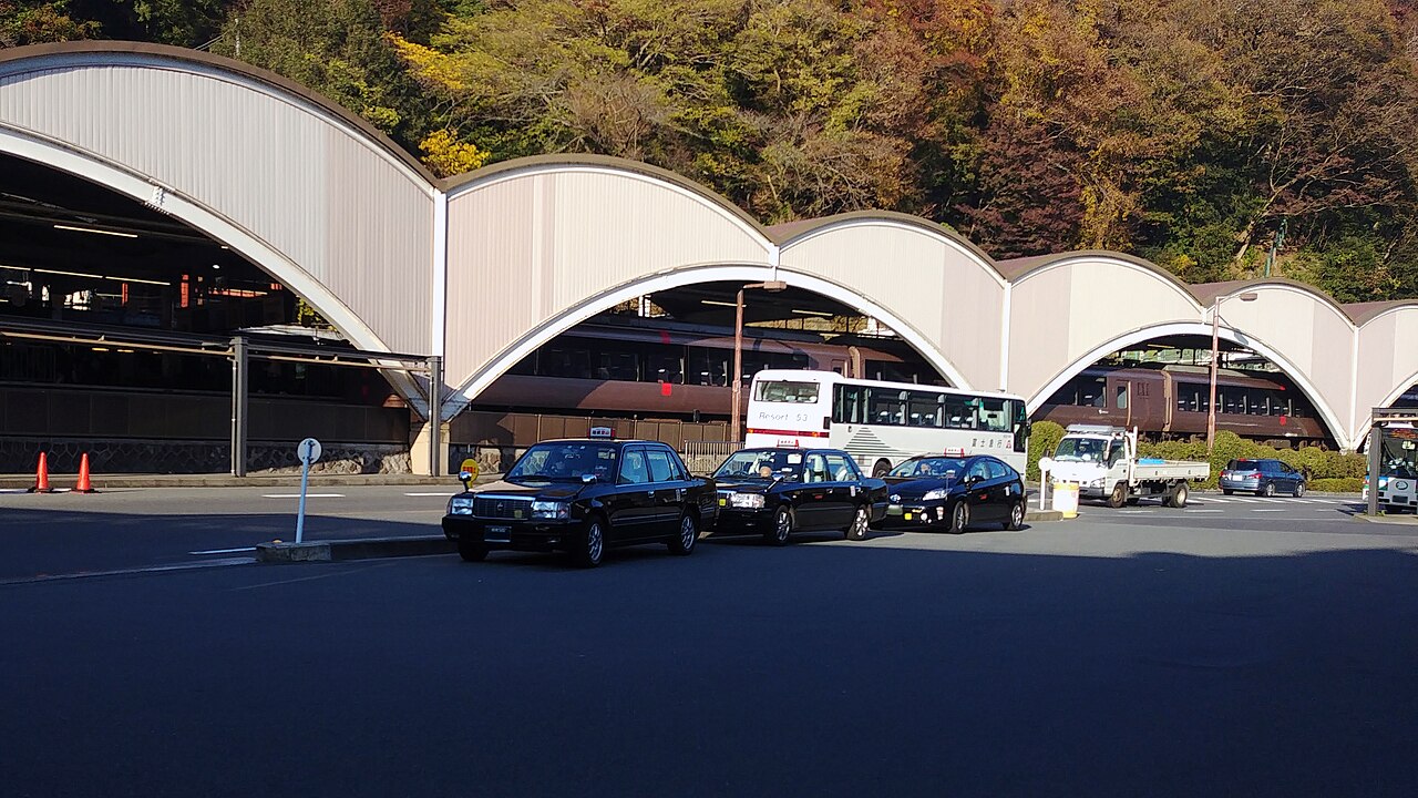 Three black taxis lined up in front of a curved-roof station building below wooded hills