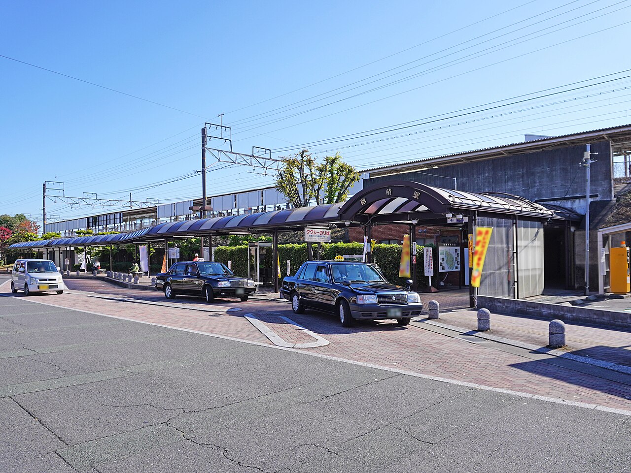 Two black taxis waiting at a small station forecourt under a curved canopy, next to a taxi stand sign