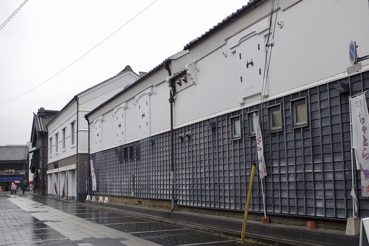 A row of large white plastered storehouses with dark tiled lower walls along a wet paved street in central Tochigi on a grey day