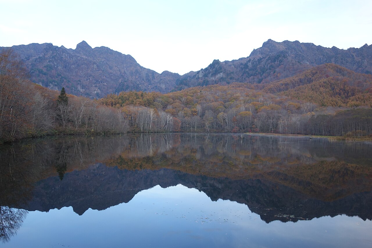 Still pond in the foreground reflecting a rugged mountain ridge and autumn-coloured slopes under a pale sky