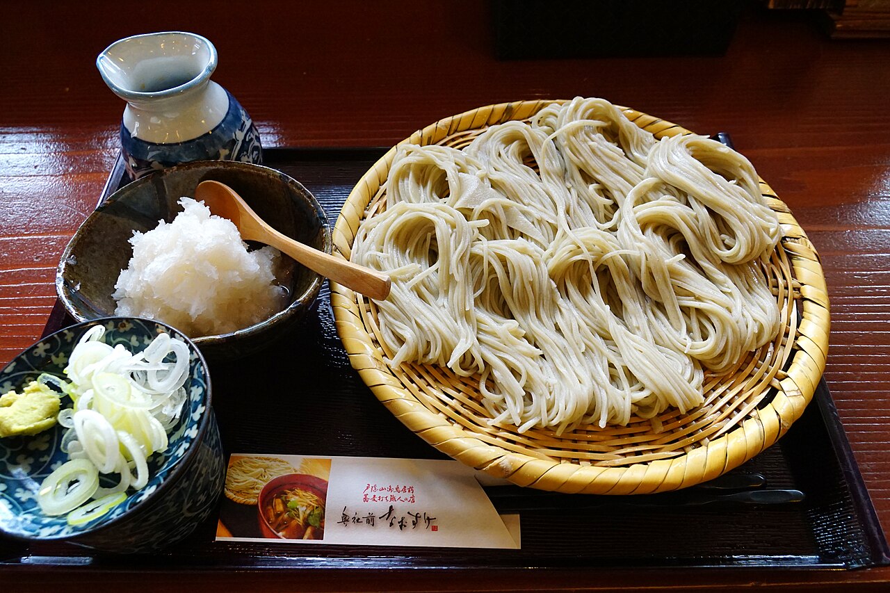 Round bamboo tray of cold soba noodles on a lacquered tray with grated daikon, chopped green onion and a small dish of dipping sauce