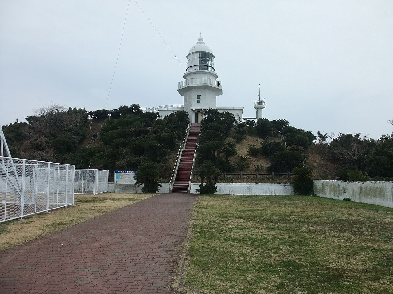 The white Toimisaki Lighthouse atop a cycad-covered rise, reached by a brick path and a red staircase