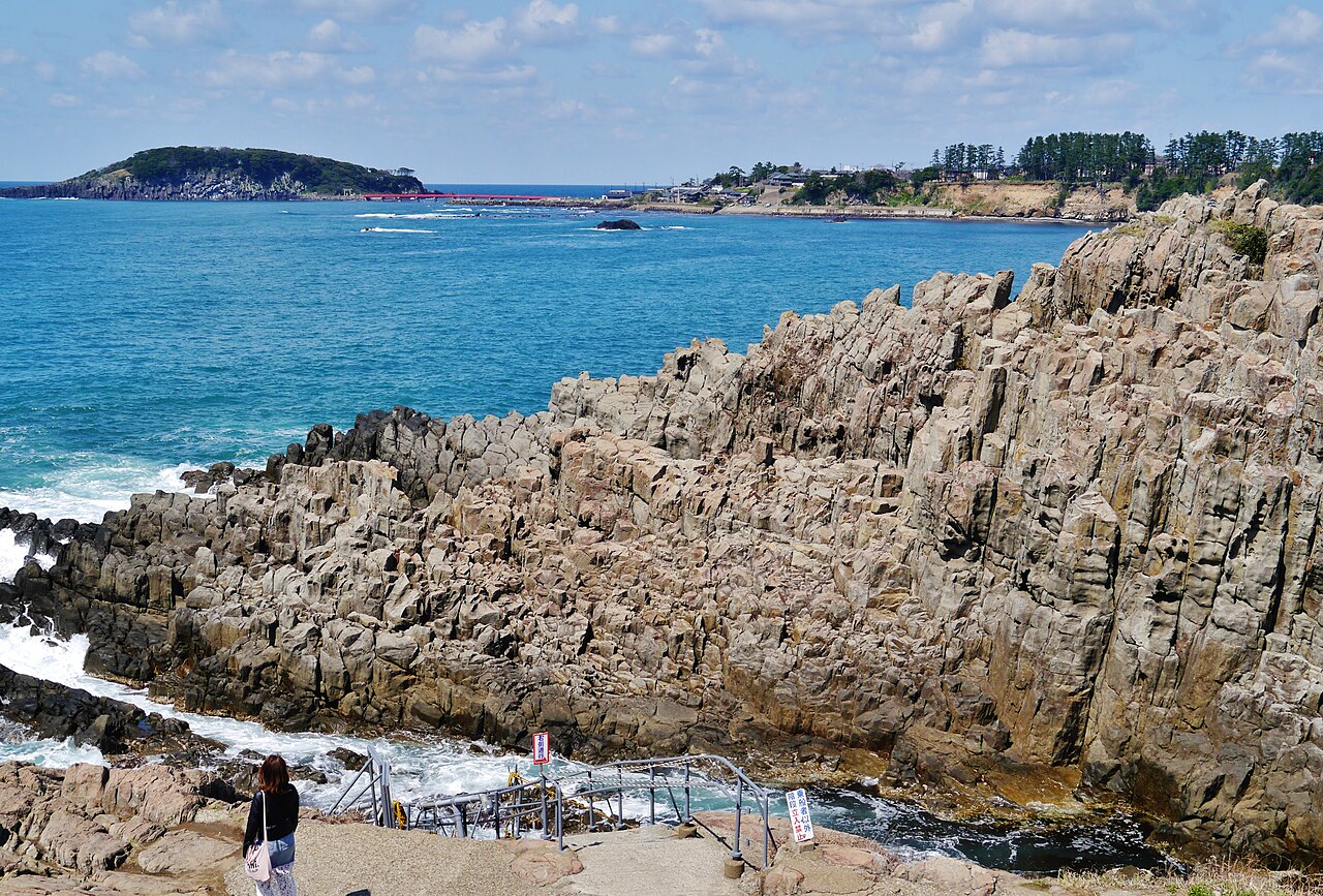 The rocky Tojinbo coastline running out toward Oshima island, with a walker on the path below the cliffs