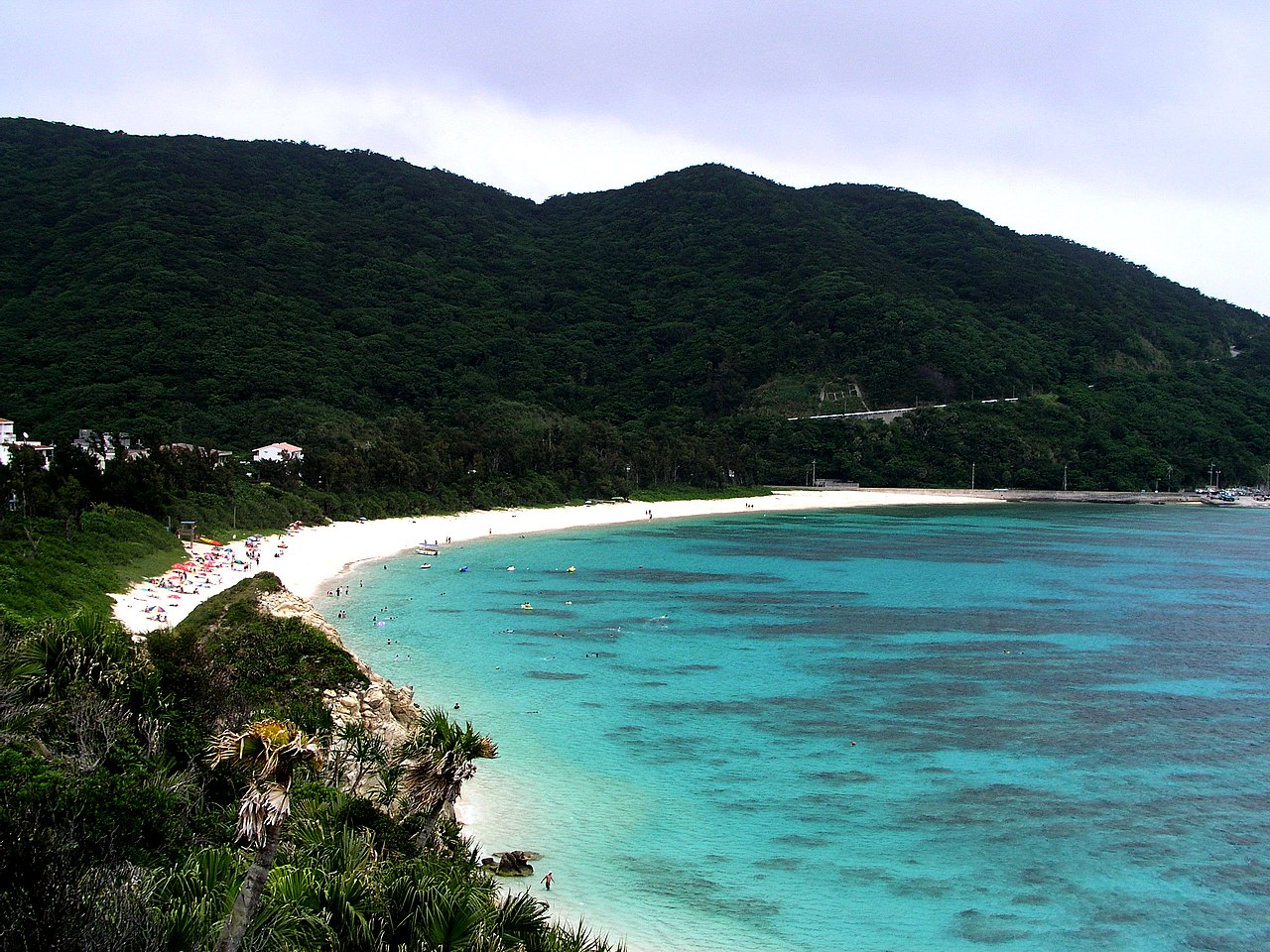 The white-sand crescent of Aharen Beach on Tokashiki Island curving along pale blue shallows, with forested hills rising behind it