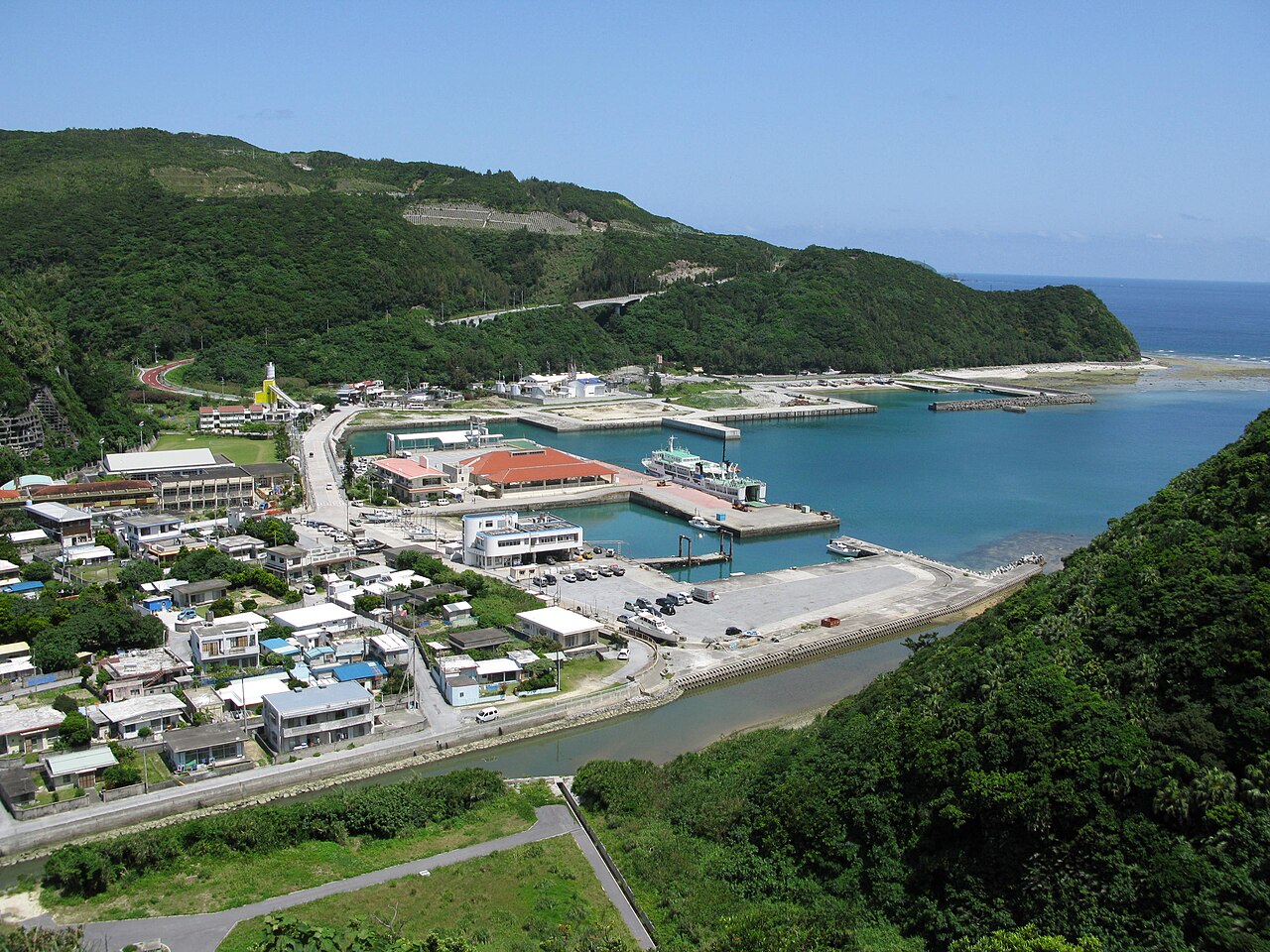 The small harbour and village of Tokashiki seen from the hillside above, with a breakwater, boats and green slopes beside the bay