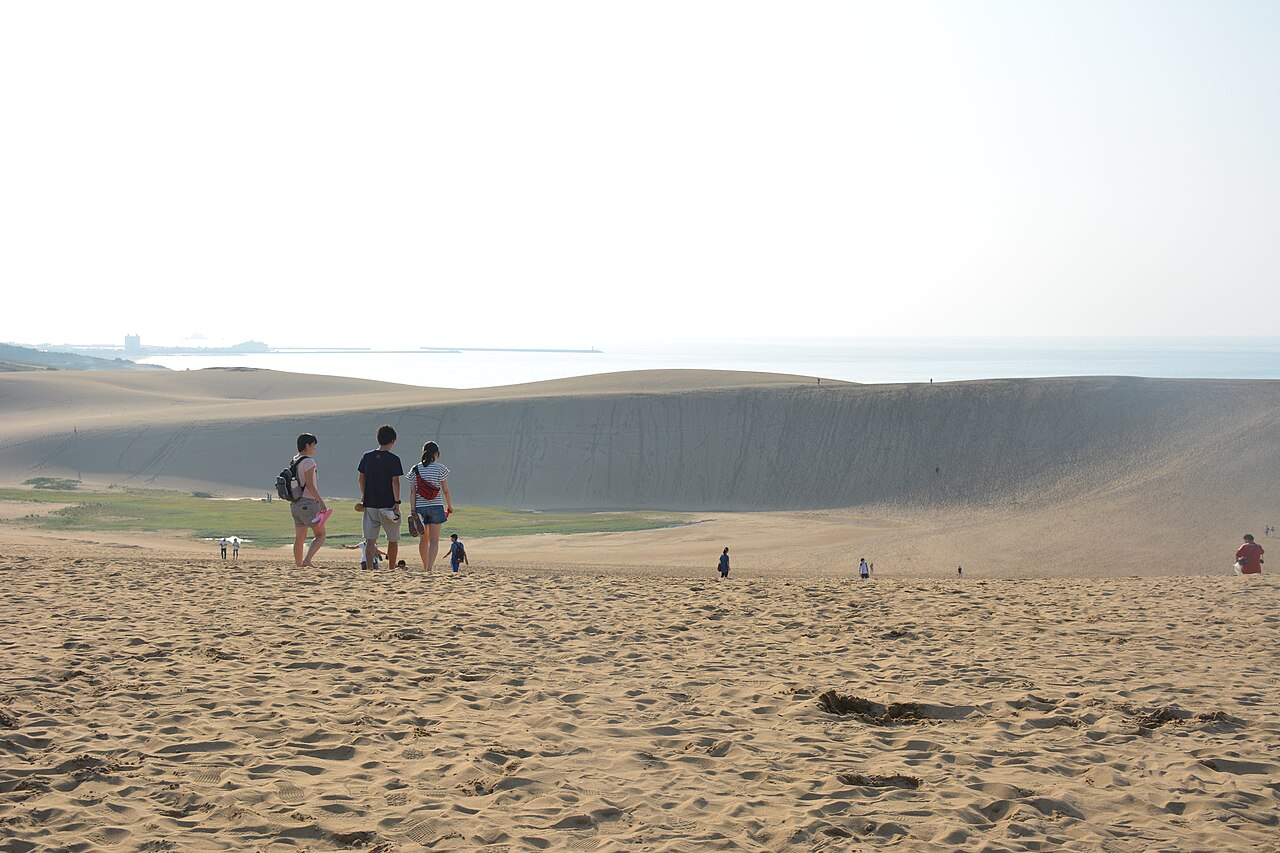 Visitors walking down a sand slope toward a long dune ridge with the Sea of Japan beyond