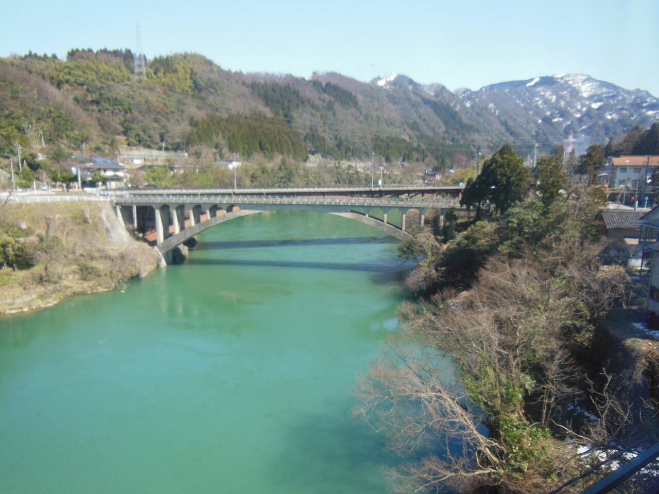 A river gorge and forested mountains seen through a train window, with a steel bridge crossing a turquoise river below.