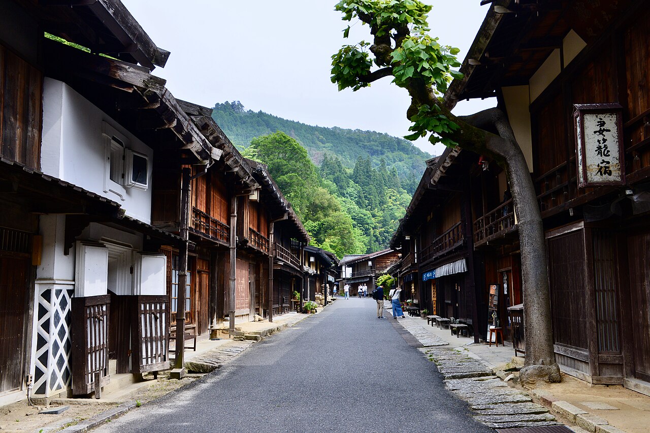 A street of dark wooden houses in Tsumago-juku, with a tree leaning over the road and forested mountains behind.