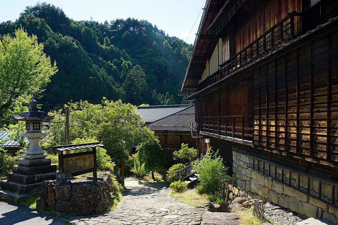 A curving stone-paved lane in Tsumago-juku lined with wooden buildings on projecting beams, a stone lantern, and forested hills behind.