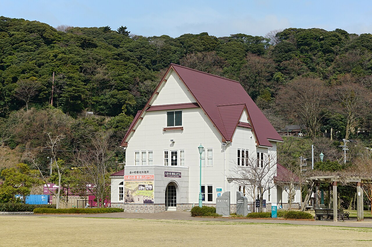 A white building with a dark red gabled roof housing the Port of Humanity Tsuruga museum, set against a wooded hillside.