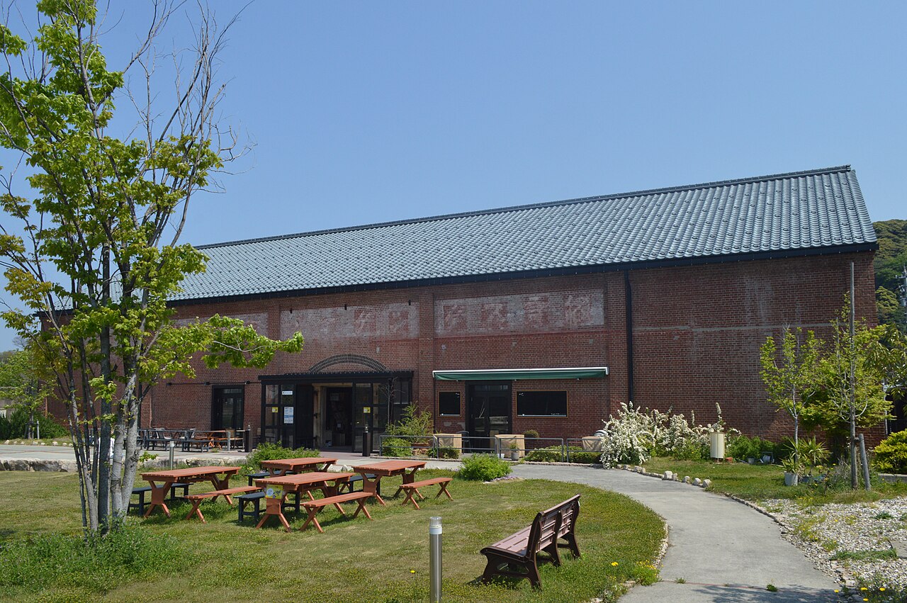 A row of red brick warehouse buildings with tiled gable roofs and arched doorways, now used as a visitor space with picnic tables outside.