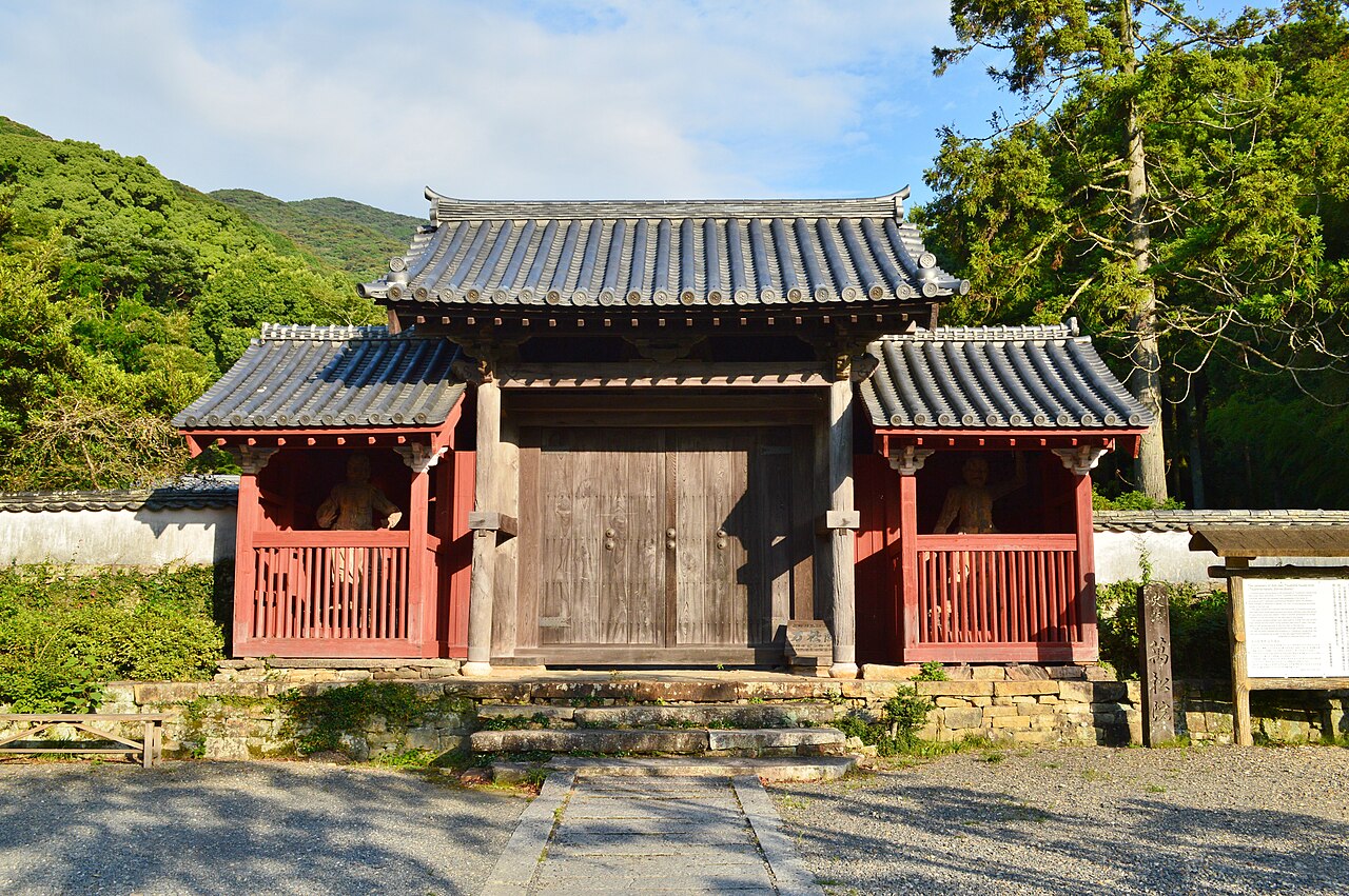 The red-pillared Sanmon gate of Banshoin temple in Izuhara, Tsushima, with a tiled roof, closed wooden doors and forested hills behind