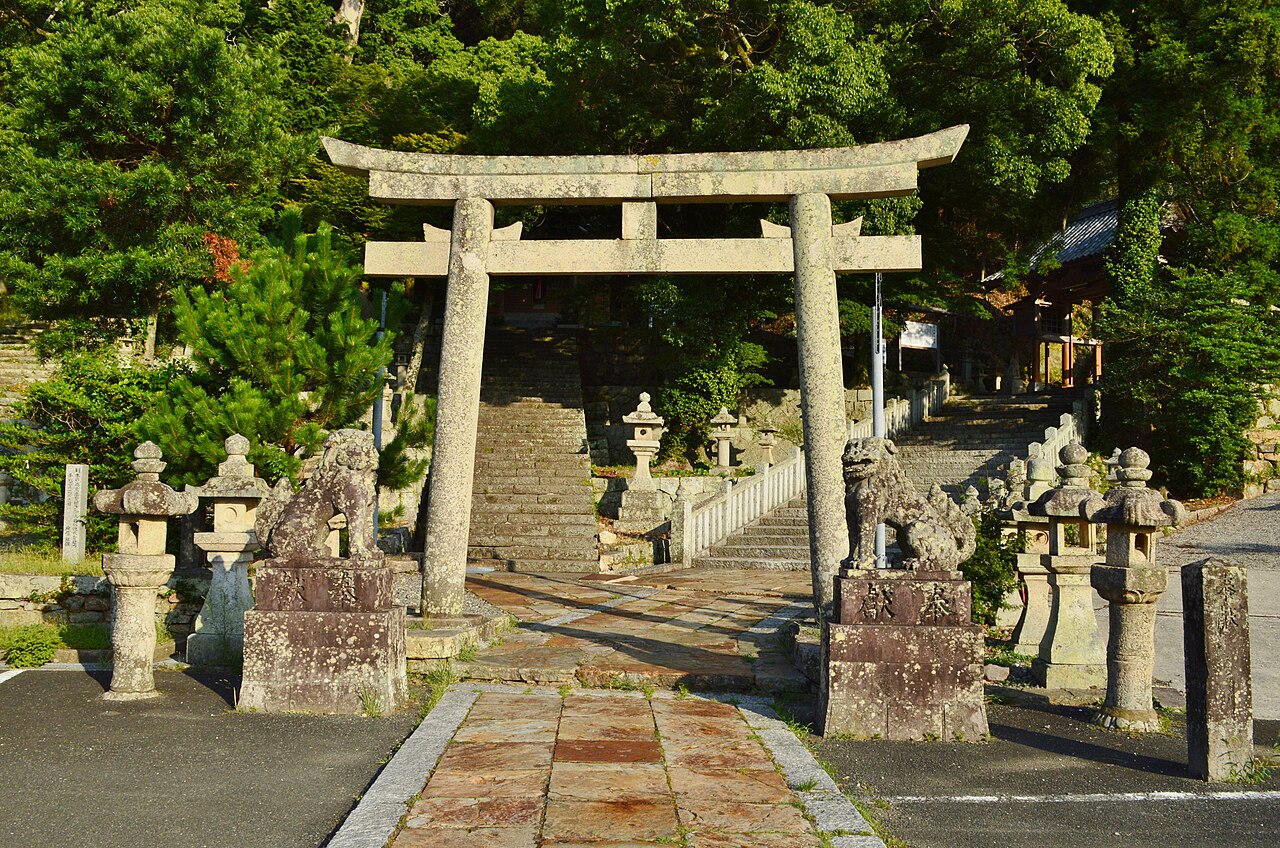 A weathered stone torii with a rope and stone guardian lions at the foot of the stairs to Izuhara Hachimangu shrine, Tsushima