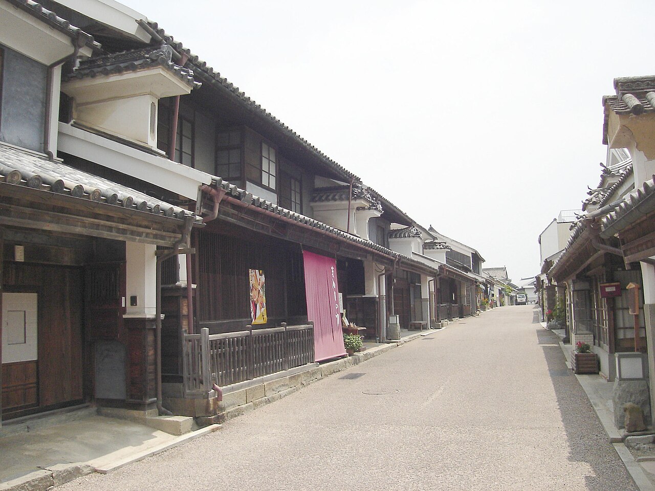 A close view of an udatsu-fronted building in Wakimachi with a lattice mushiko window and a pink cloth banner beside the entrance, along a street lined with more such houses.