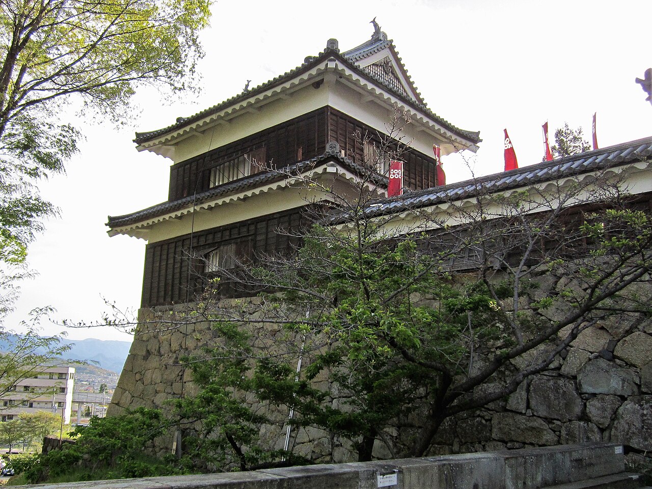 The two-storey South Turret of Ueda Castle rising from a rough stone wall, with red Sanada banners above the white-trimmed eaves