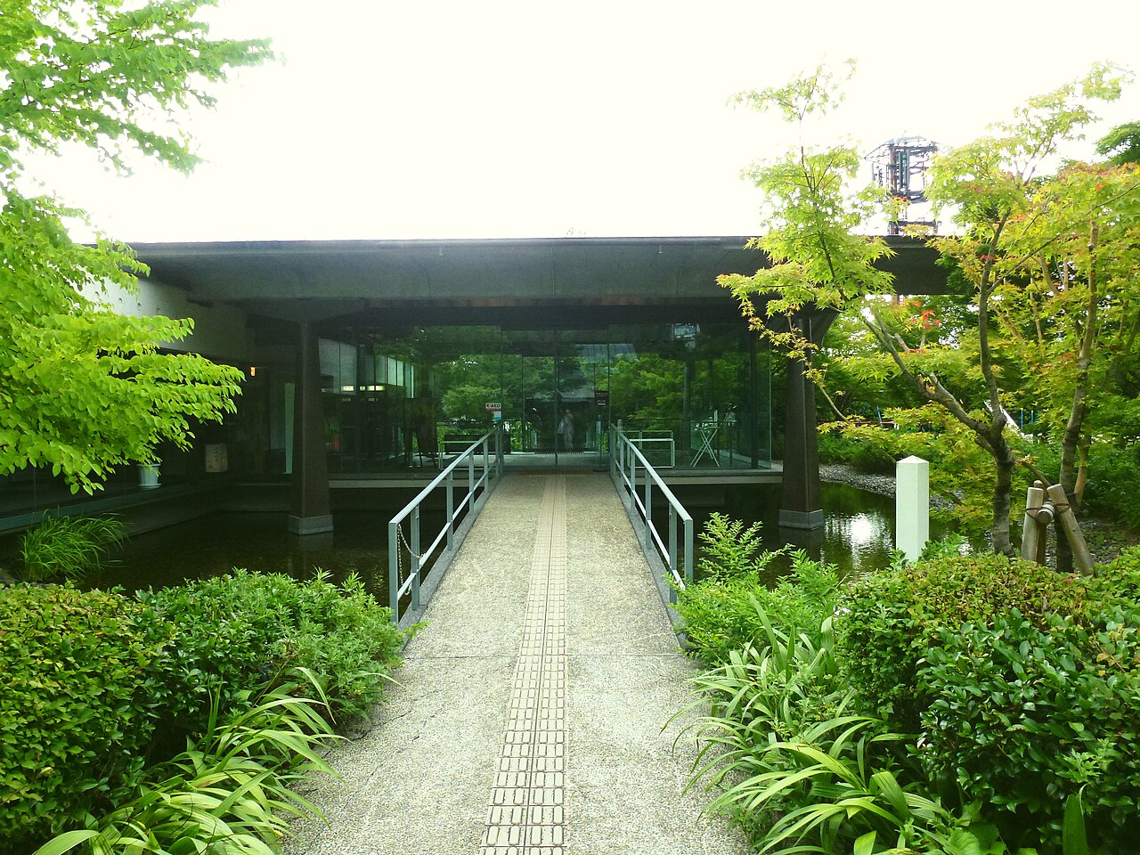A low glass building with a walkway over water leading to the entrance of the Tale of Genji Museum