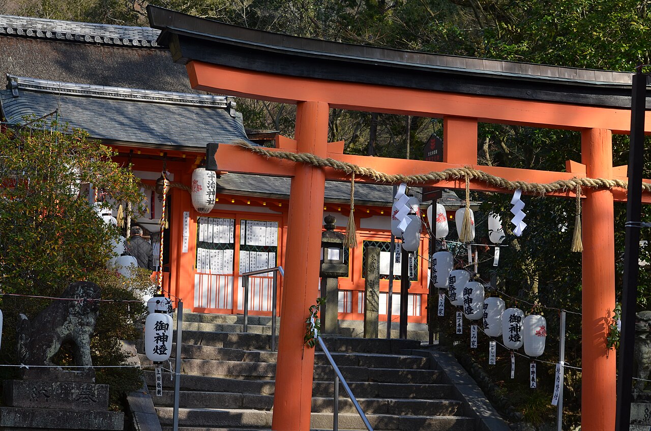 A vermilion torii with a shimenawa rope and white paper lanterns at the foot of stone steps at Uji Shrine