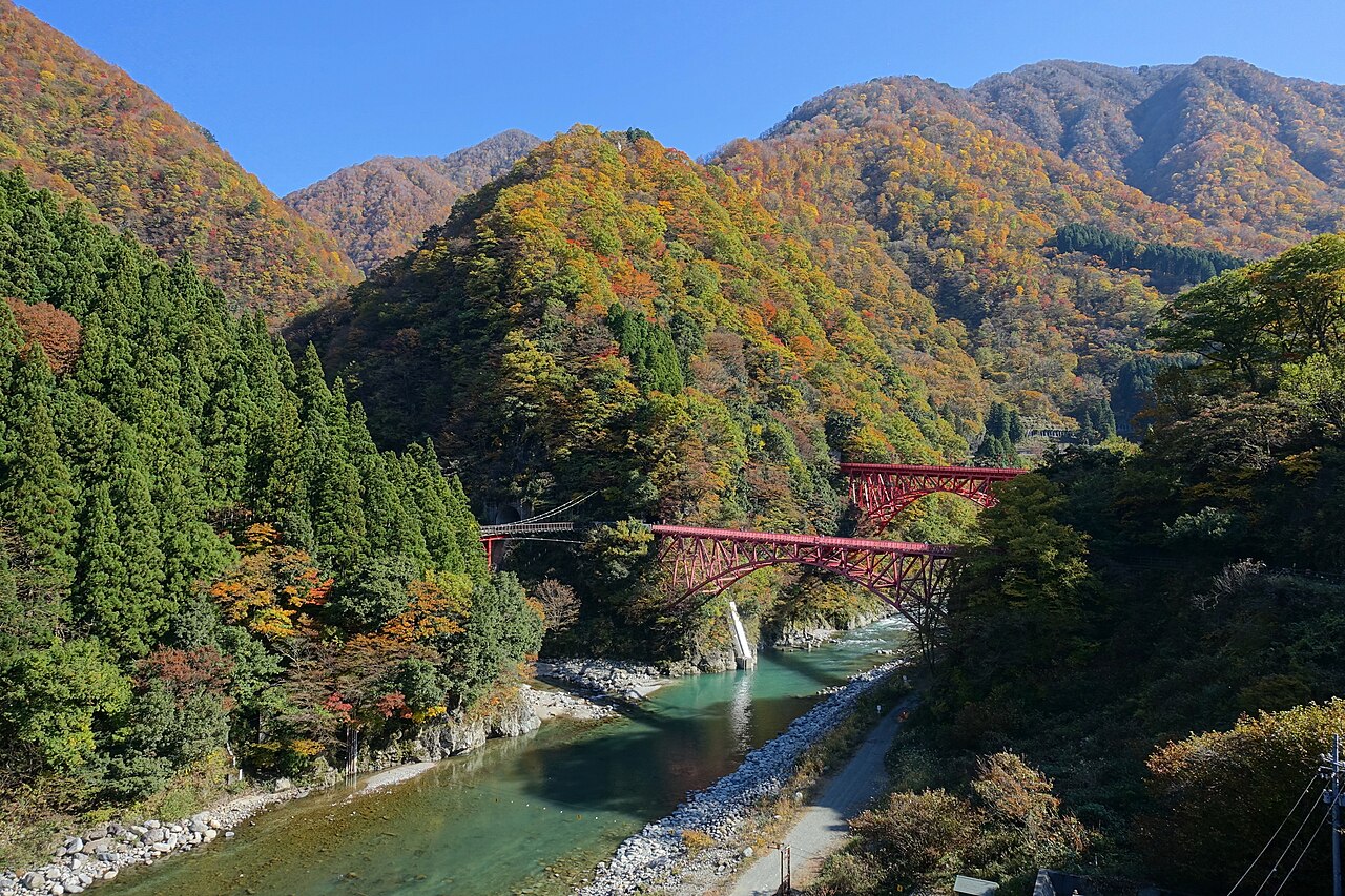 Two red steel railway bridges span a turquoise river in a deep Kurobe Gorge valley, with autumn-coloured forest on the hillsides