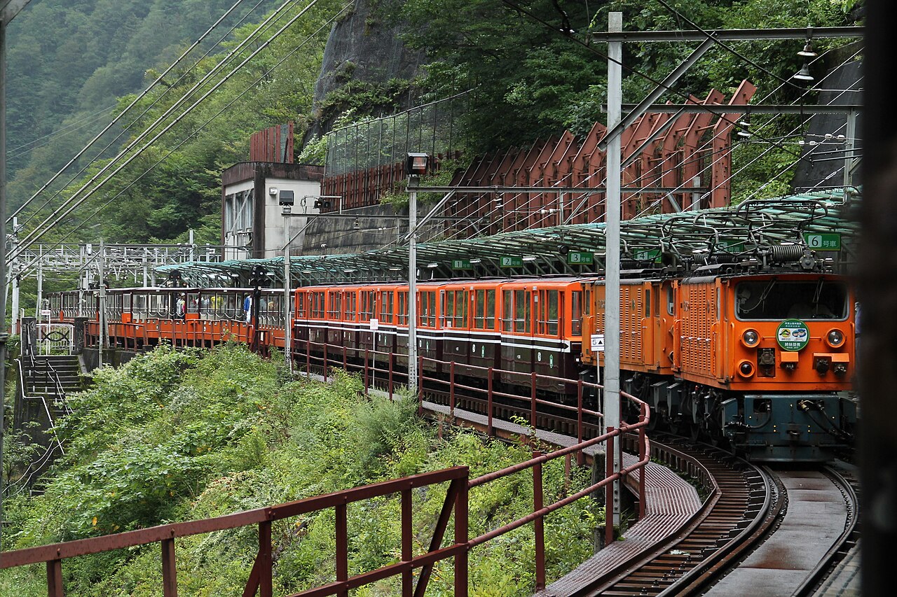 An orange Kurobe Gorge Railway locomotive with open-sided passenger cars pulls under a covered platform among green hills
