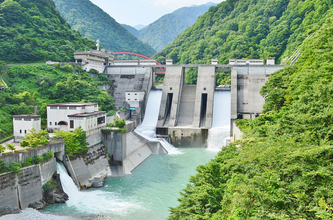 Water pours from the spillway gates of Unazuki Dam into a turquoise pool, with a power station building and green mountainsides around it