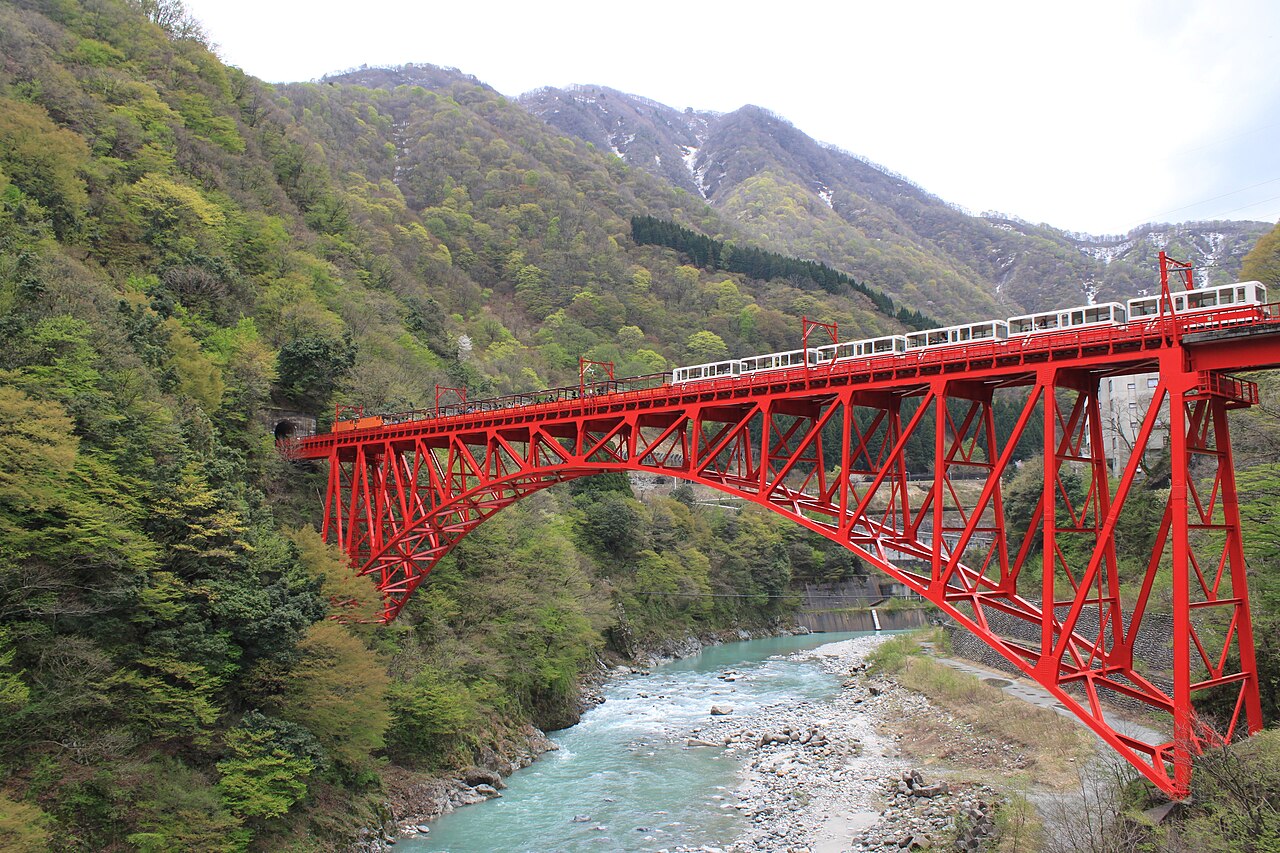 A white Kurobe Gorge Railway locomotive and cars cross a tall red arch bridge above a river, with snow-streaked mountains in the distance