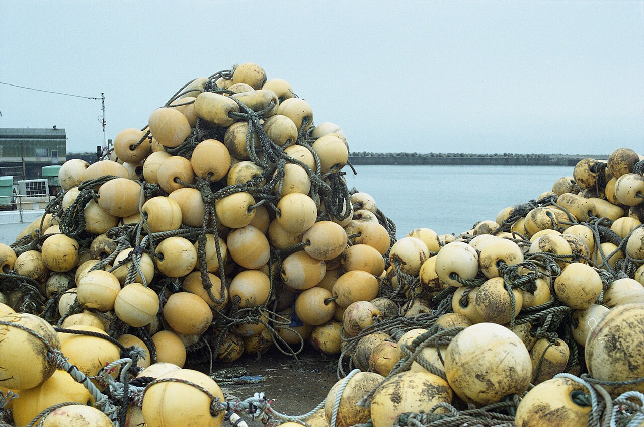 A heap of yellow fishing-net floats tangled in rope on a quay, with calm harbor water and a long breakwater behind.