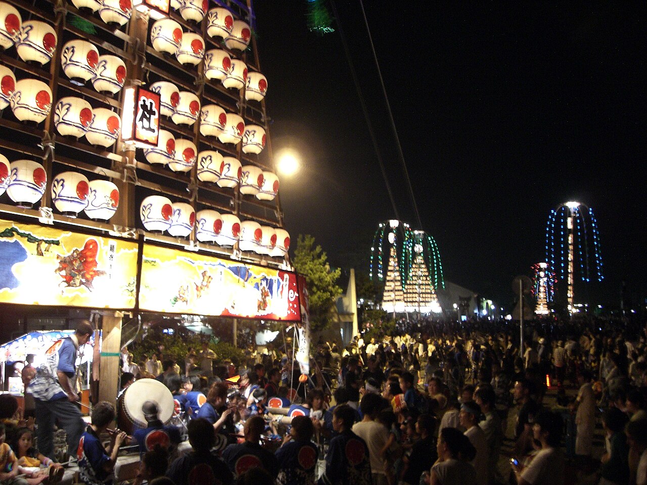 A night festival crowd in front of a tall wall of lit paper lanterns on a float, with more illuminated floats glowing in the background.