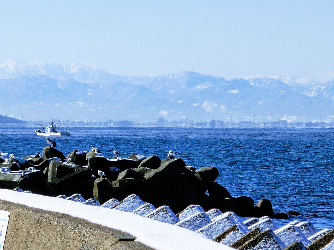 Deep blue sea behind a row of concrete breakwater blocks, with a long snow-dusted mountain range and a faint far shore under a clear sky.