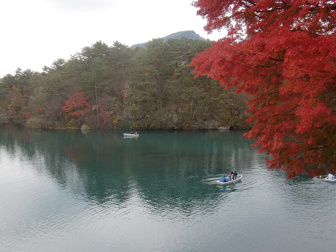 Rowboats on the teal water of Bishamon-numa at Goshikinuma in autumn, with a bright red maple branch in the foreground and a pine-covered shore and mountain beyond.