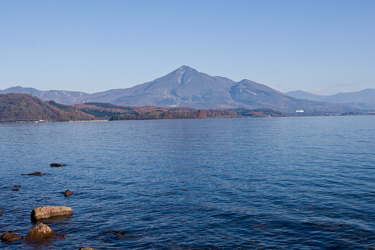Mount Bandai rising behind the blue water of Lake Inawashiro, with a few rocks in the shallows in the foreground.