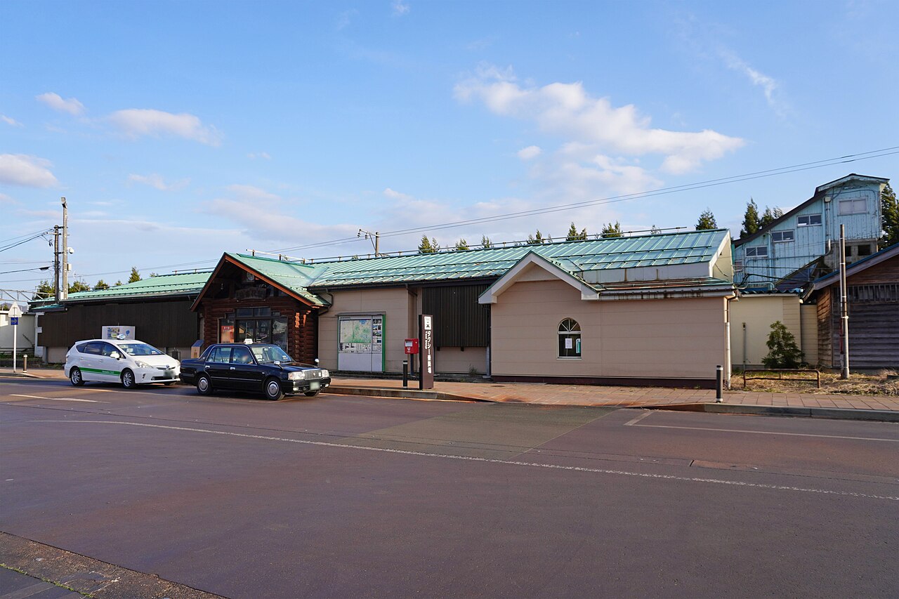 The log-style front of the JR Inawashiro railway building with a green roof and cars parked in front, under a blue sky.