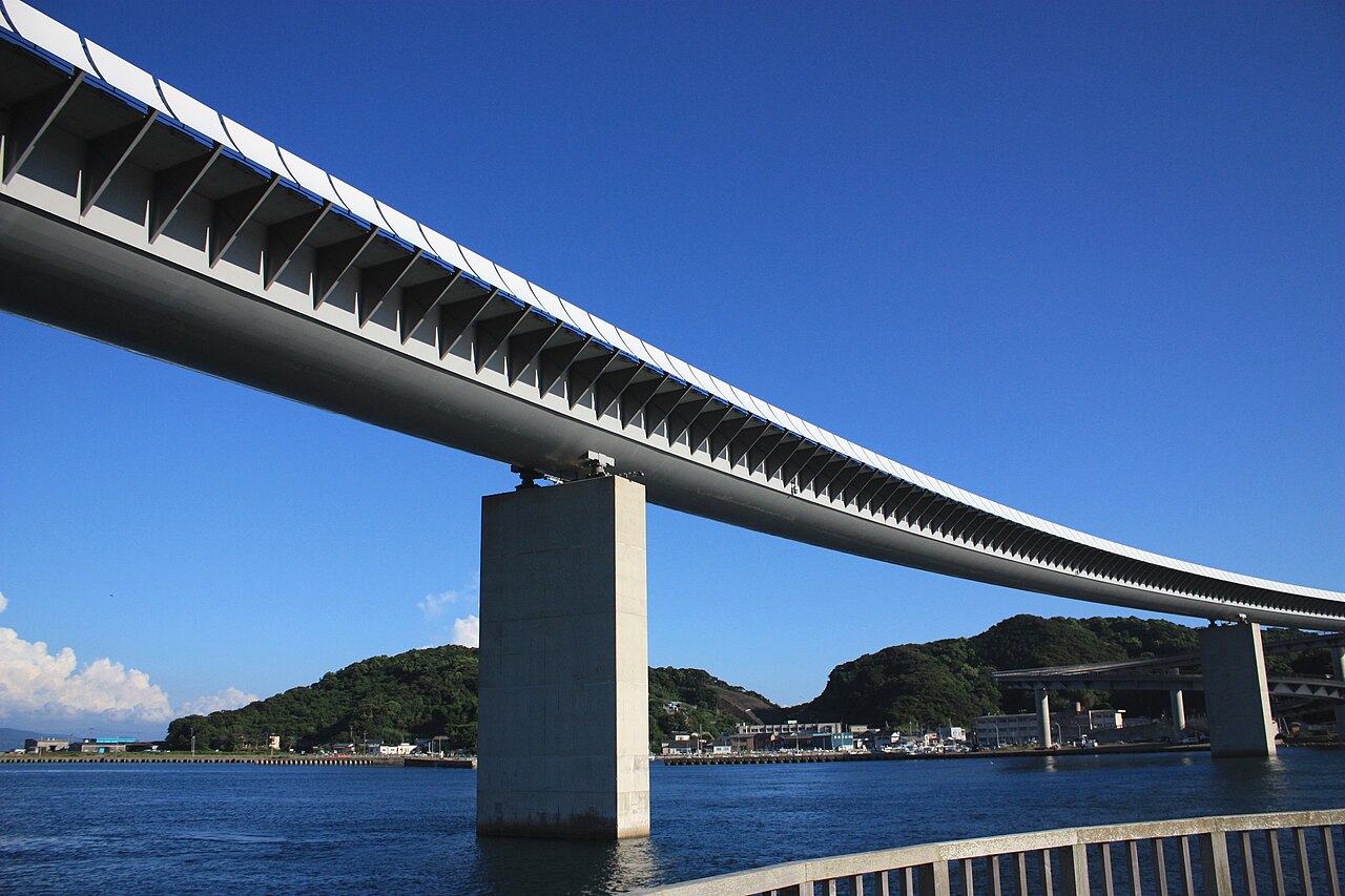 The long white underside of a curving road bridge crossing a harbor channel under a deep blue sky, with a concrete pier standing in the water and green hills beyond