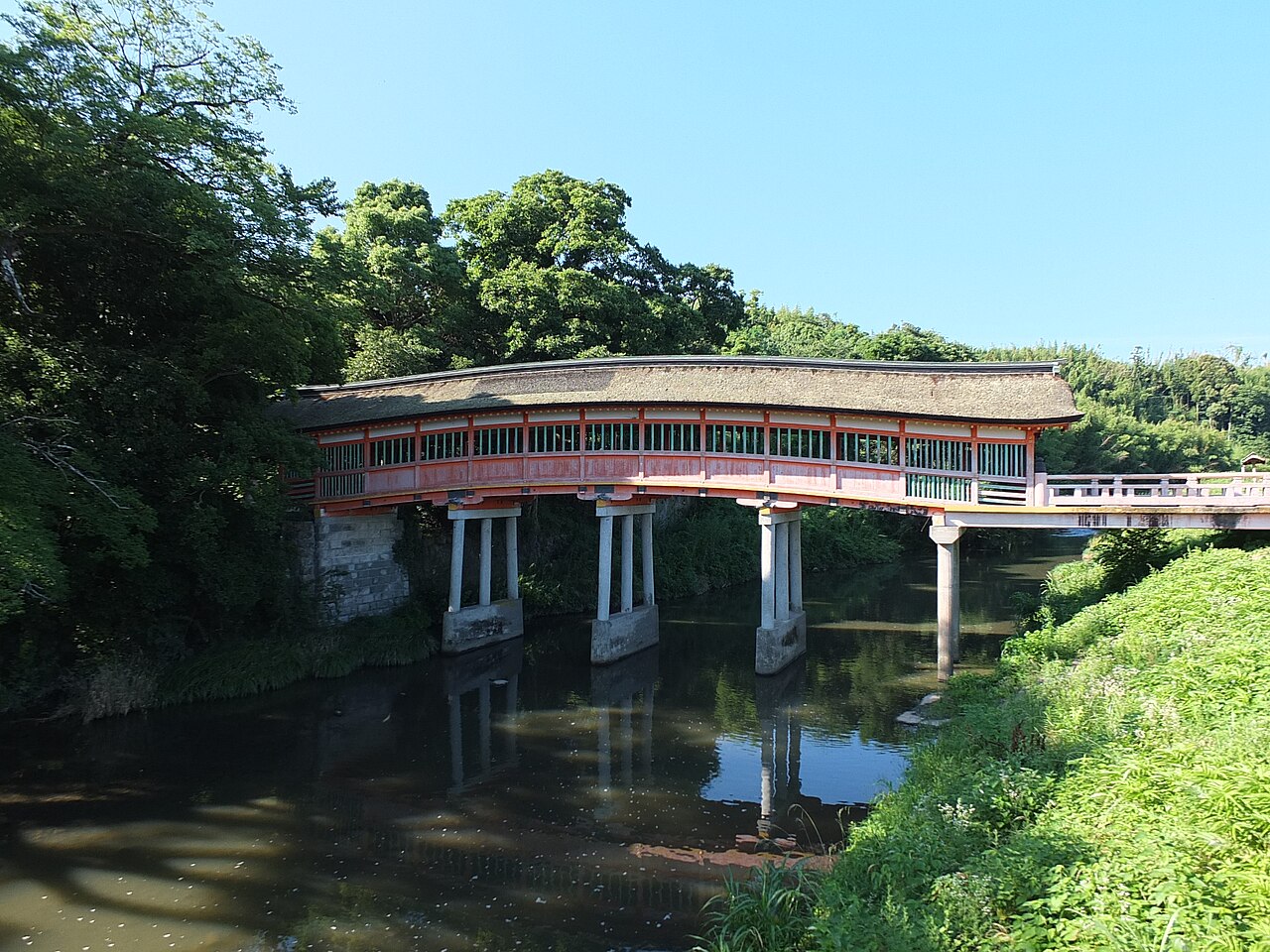 A long covered bridge with a thatched curved roof and vermilion rail, resting on pale stone piers over a calm river that mirrors the green trees, under a clear blue sky.