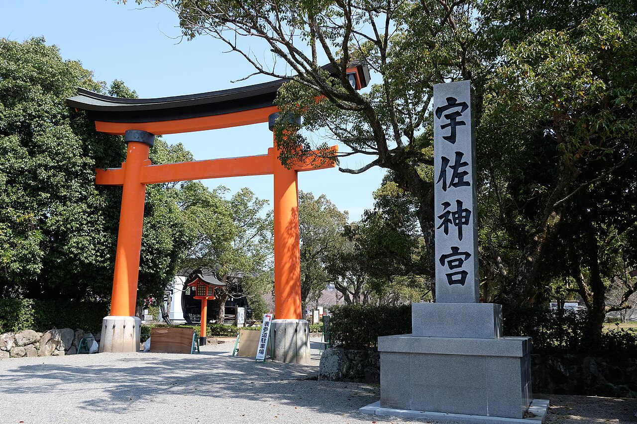 A large vermilion torii gate with a curved dark top beam beside a tall grey stone pillar carrying four large black characters, with green trees behind and a gravel forecourt in front.