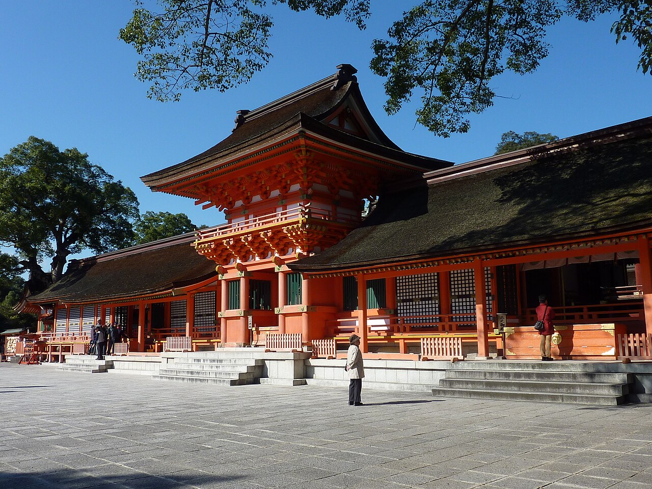 A two-storey vermilion gate with a curved dark thatched roof and gold fittings beside long vermilion halls, seen across a wide paved courtyard under a clear blue sky with a few small visitors and large trees at the left.