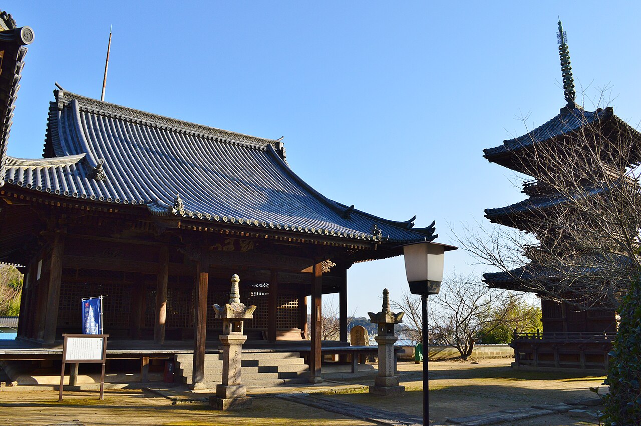 The main hall of Honren-ji in Ushimado, with a sweeping blue tiled roof, stone lanterns in the courtyard and the three-storey pagoda at the right edge