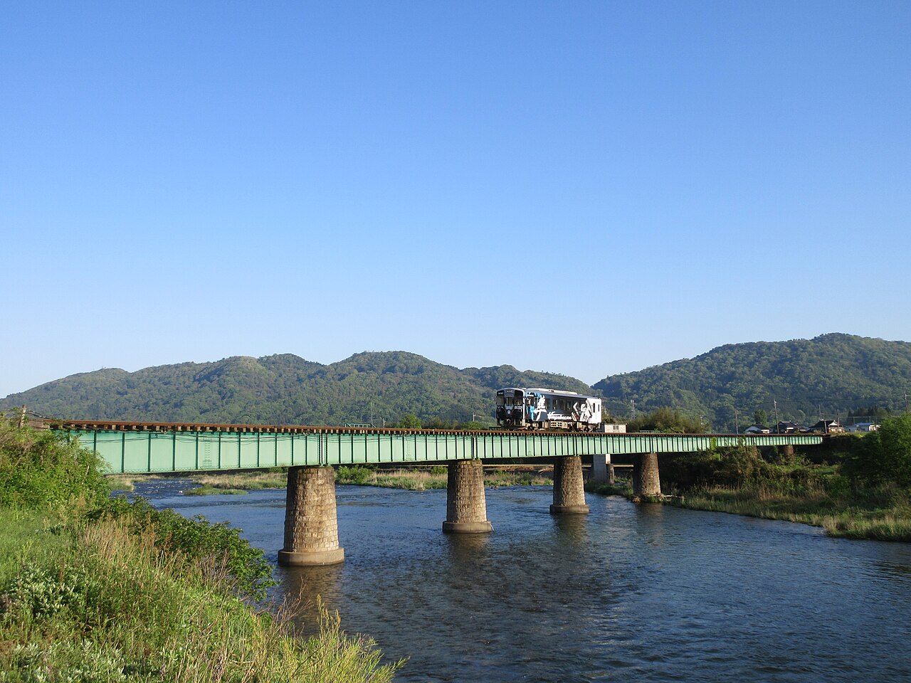 A single Wakasa Railway railcar crossing the green steel No.1 Hatto River Bridge on stone piers, with green hills behind