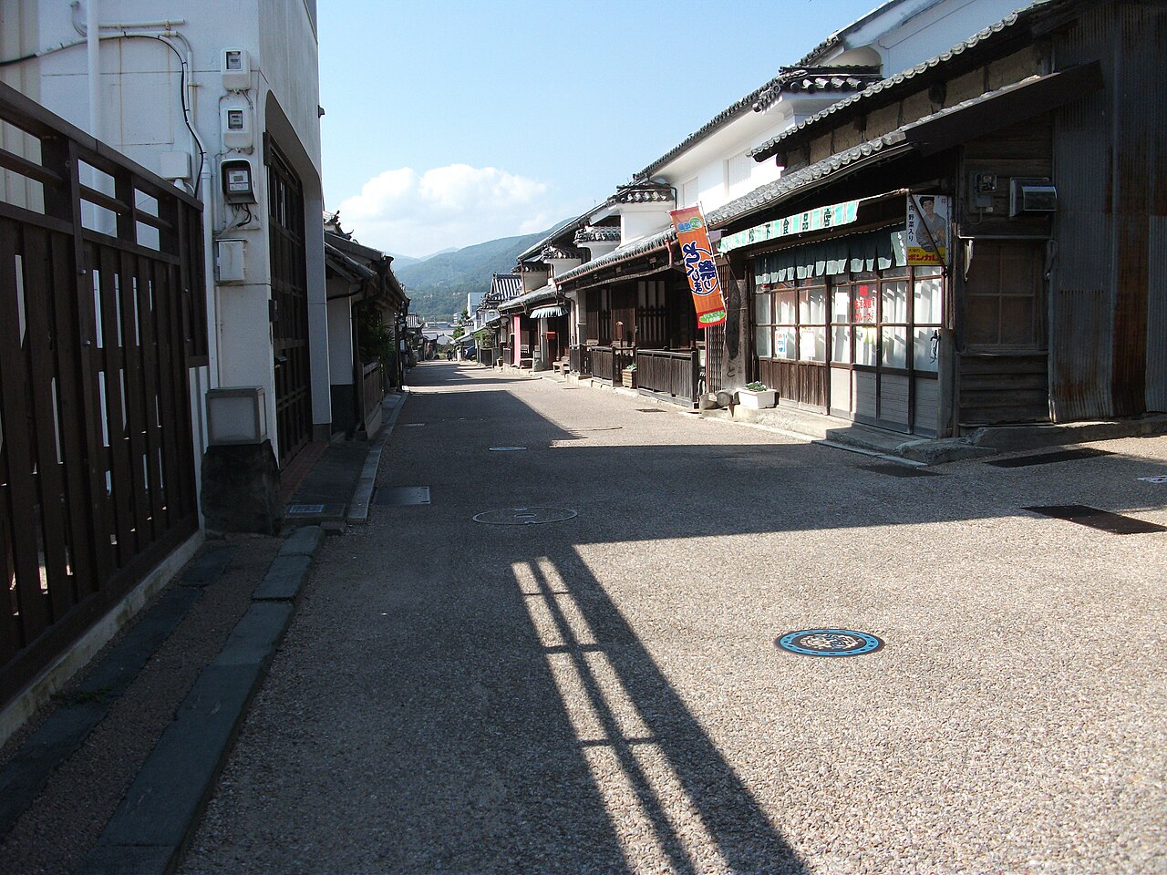 A street in Wakimachi lined with tiled-roof shops and udatsu walls, with an orange banner hanging in front of one storefront under a clear sky.