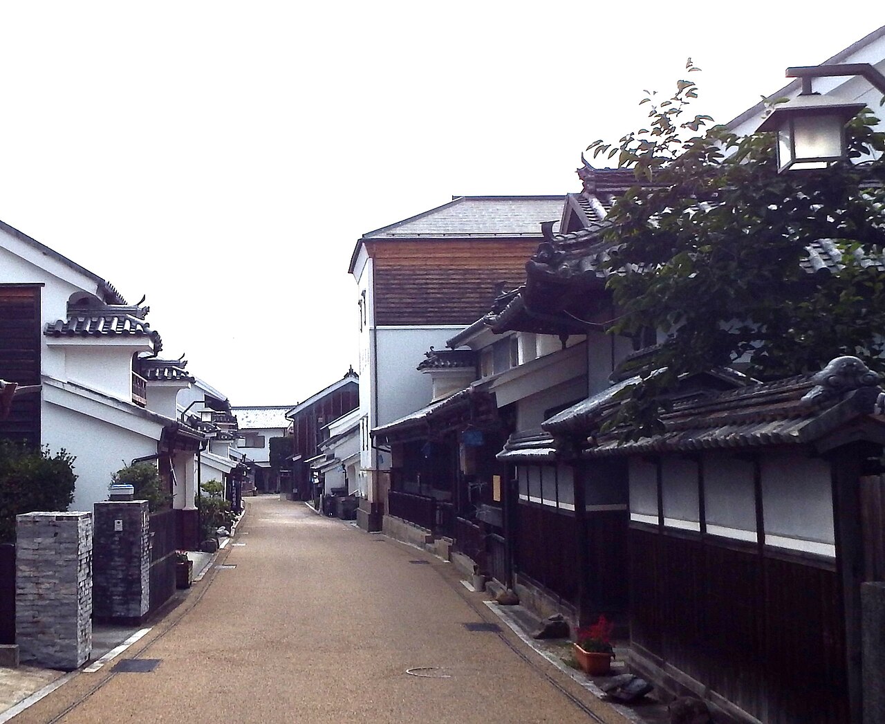 A view along a paved lane in Wakimachi flanked by whitewashed udatsu houses, with a tree's green branches leaning over the street in the foreground.